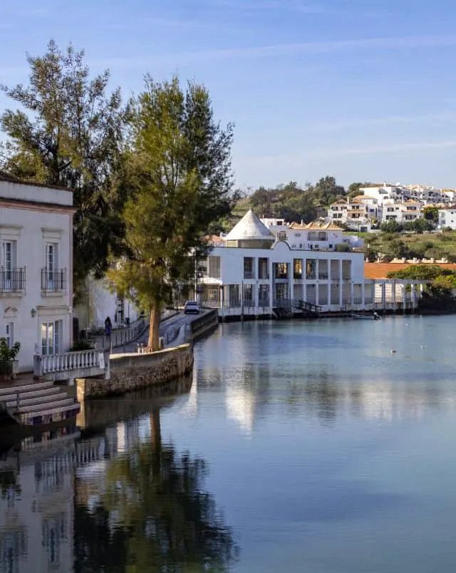 fun activities in Tavira, Two storey residential buildings sitting on the edge of a body of water with rolling hills behind all under a blue sky with slight mist