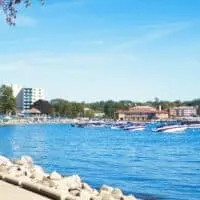 View of coastal harbour with several speedboats moored in front of people sunbathing on the beach with green trees behind on a bright sunny day