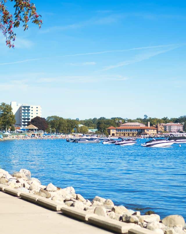 Lake Geneva Resorts for families, View of coastal harbour with several speedboats moored in front of people sunbathing on the beach with green trees behind on a bright sunny day