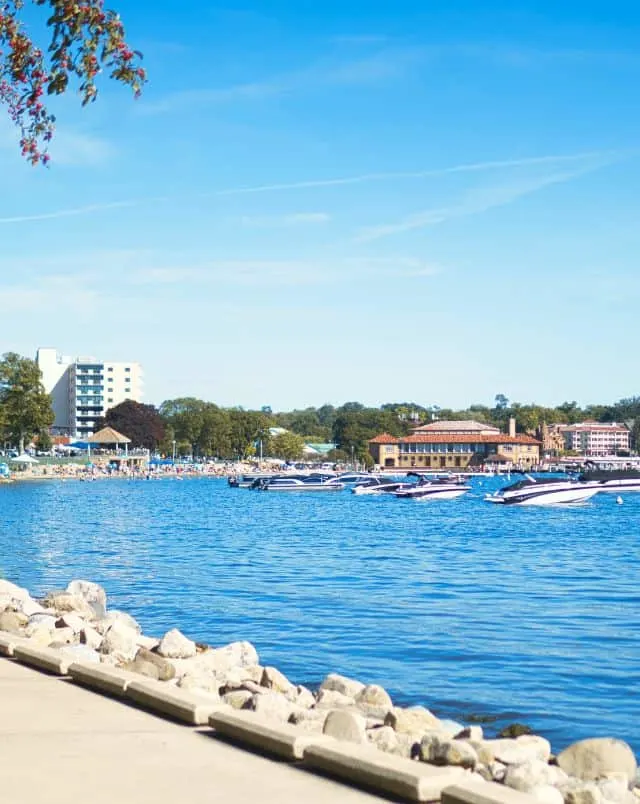 View of coastal harbour with several speedboats moored in front of people sunbathing on the beach with green trees behind on a bright sunny day