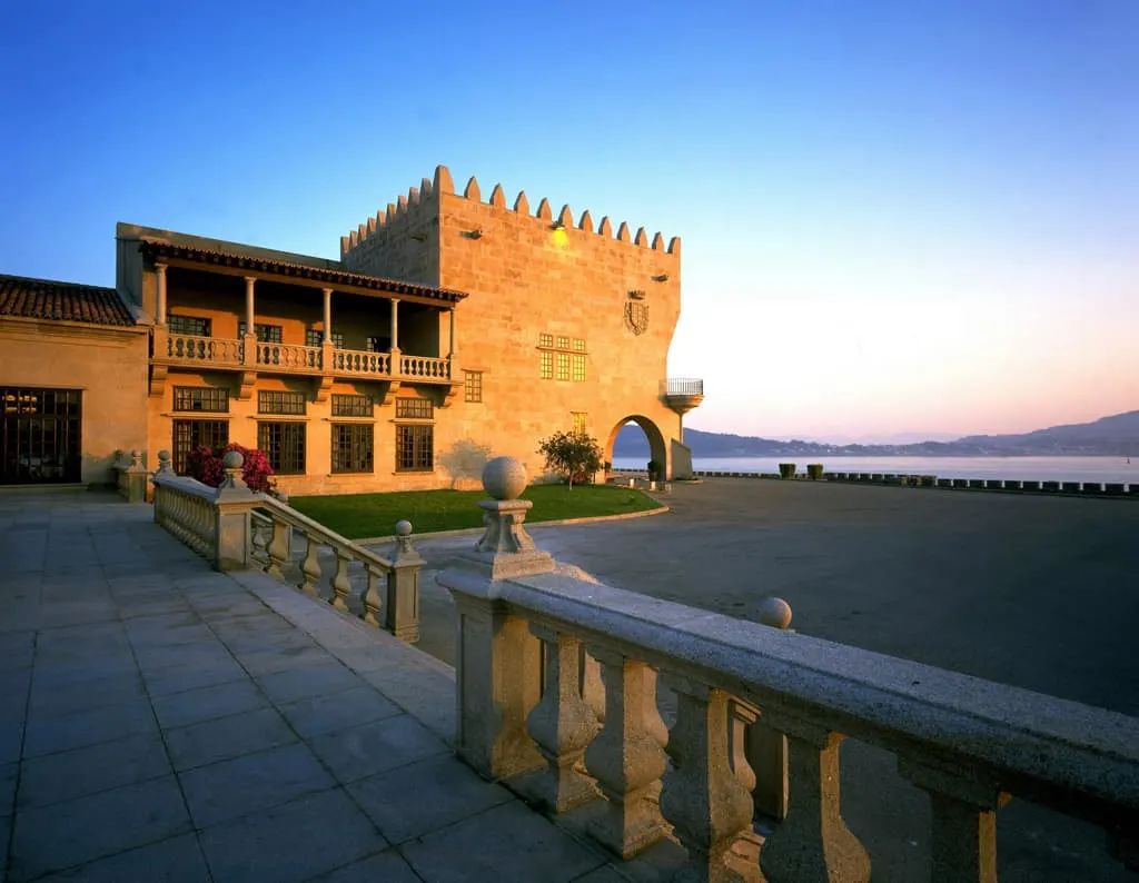 Stay in a spanish parador this summer, view of outdoor stone courtyard next to the coast with ornate stone structure lit by the orange rays of the setting sun
