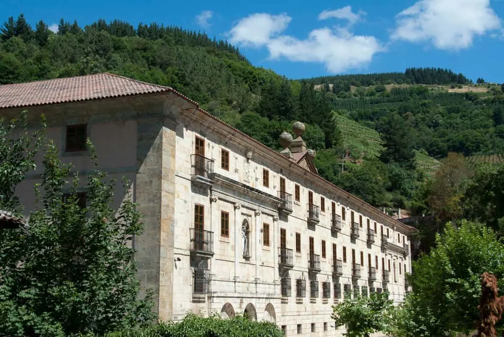 Stay in some of the most luxurious parador hotels northern spain has to offer, view of Parador de Corias with long stone front lined with many square windows set into the side of a large green hill with rolling fields and forests behind