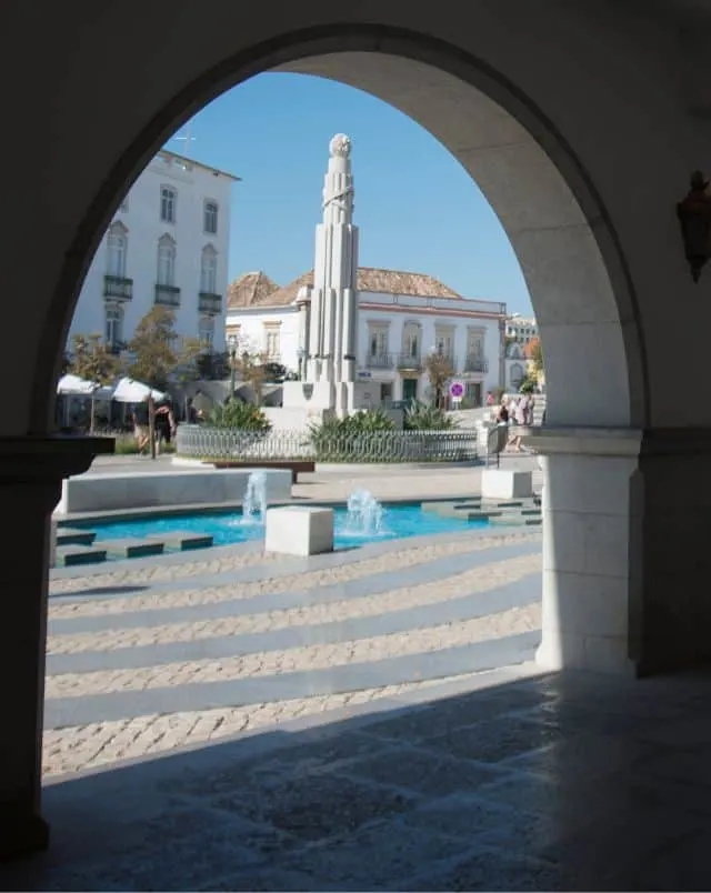 Tavira things to do, View through a stone arch of an outdoor square with pools and fountains as well as a tall stone statue with other stone buildings behind on a bright clear day