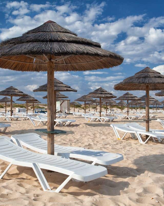 best beaches in Tavira Portugal, A multitude of sun loungers sitting in sand under wooden beach umbrellas all under a blue sky with white clouds
