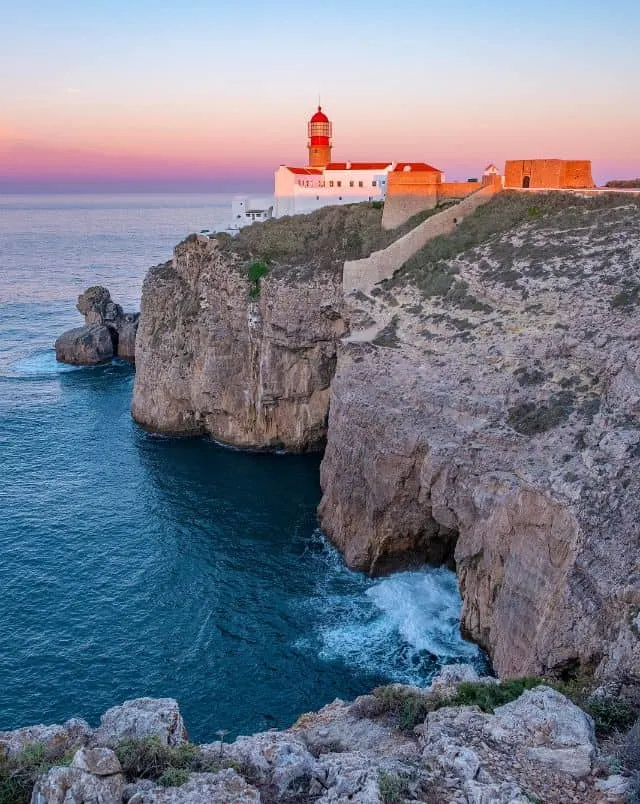 best places to stay in Algarve, Rocky cliffs on the shores of the ocean with old fortifications and a more modern lighthouse resting on the edge lit by the orange rays of the setting sun under a clear dusk sky in sagres