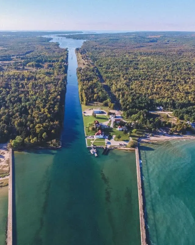 best coastal towns in Wisconsin, Aerial view of green forest area with small open area containing a few buildings sitting on the coast next to the turquoise waters of the sea and a long river stretching off into the distance