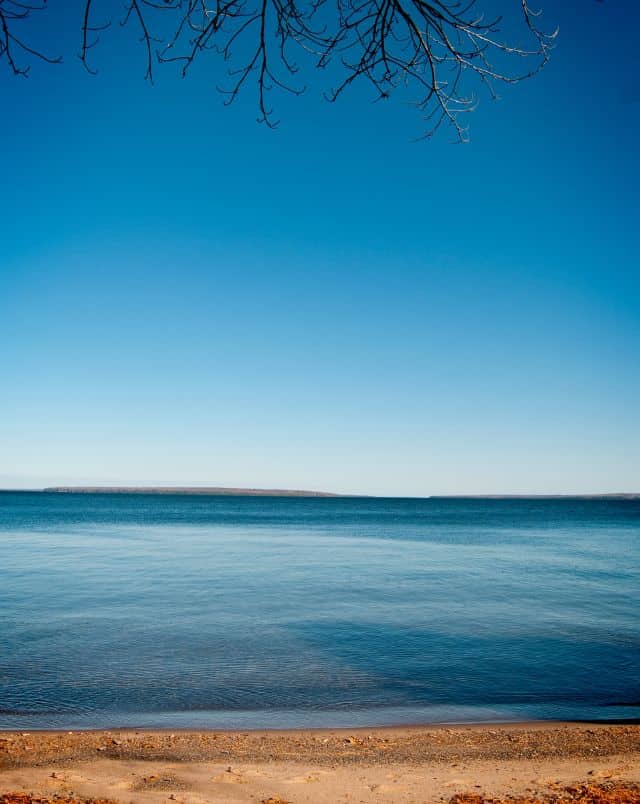 Best Lake Superior towns, View looking out to sea with sandy beach and tree branches in the foreground and the deep blue sea beyond under a wide open blue sky