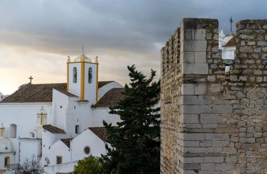 nicest towns in Algarve Portugal, Stone castle tower fortification with large white church behind under an ominous grey cloudy sky at sunset