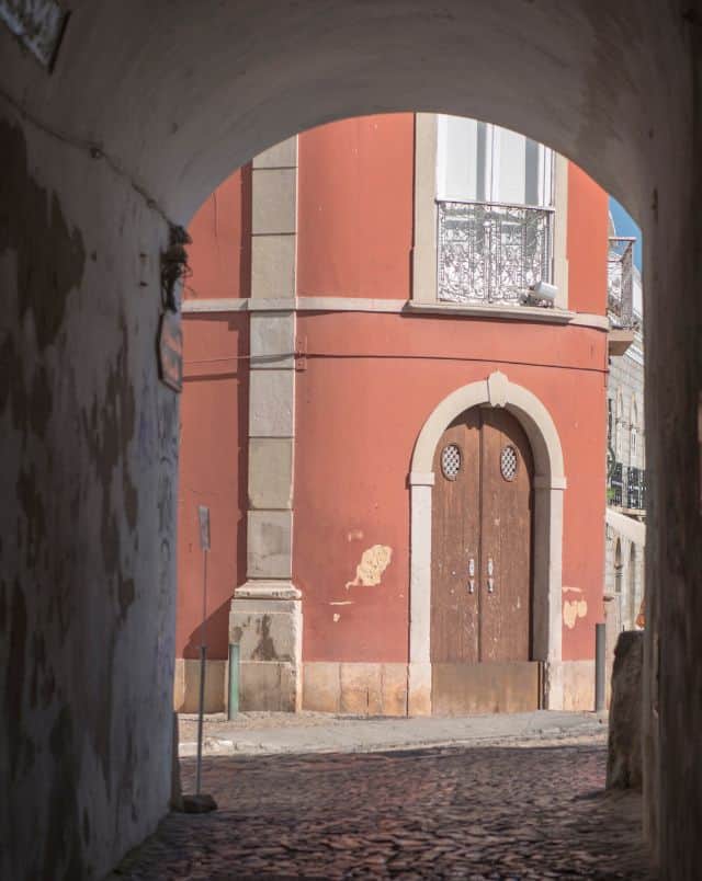 what to see in Tavira Portugal, View through an archway covering a cobbled street of a tall peach-cloured old residential building with arched wooden door