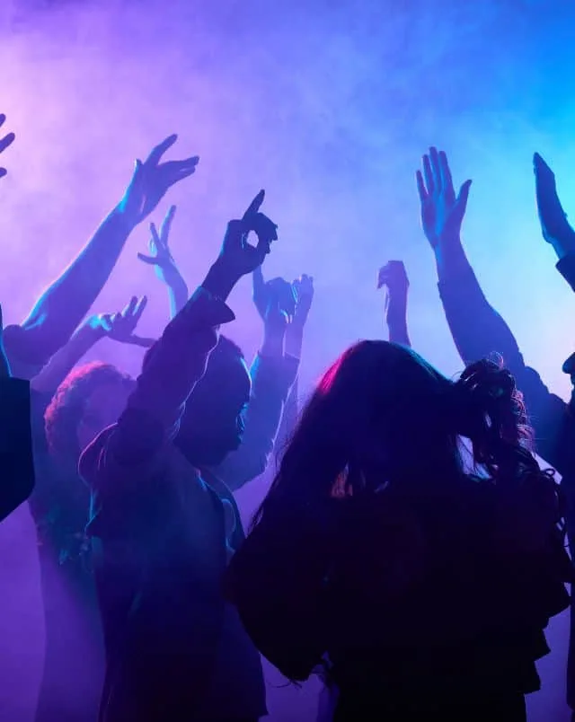 Several people in a club raising their hands as purple light reflects off of smoke swirling around them