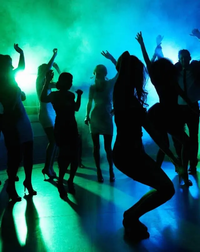 View of silhouettes of many people dancing on a dance floor in a club surrounded by smoke and lit by green and blue neon lights