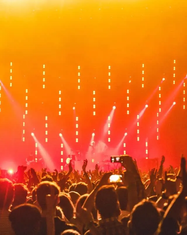 View over the top of a large group of people dancing on a dance floor in a club towards a stage with musicians lit by spotlights all surrounded by a wall of red and orange lights