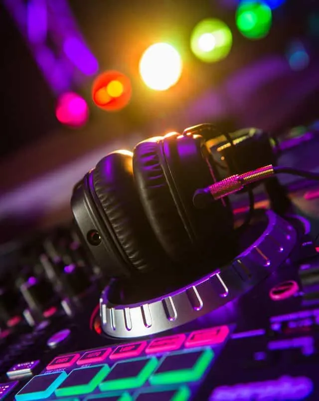 Close up shot of a pair of headphones resting on top of a DJ's mixing desk with bright rainbow coloured lights behind