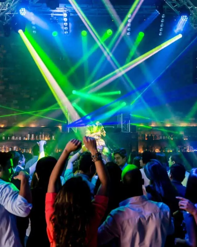 Interior of club with a crowd of people dancing under neon green and blue strobe lights and spotlights with lighting rigs suspended from the ceiling