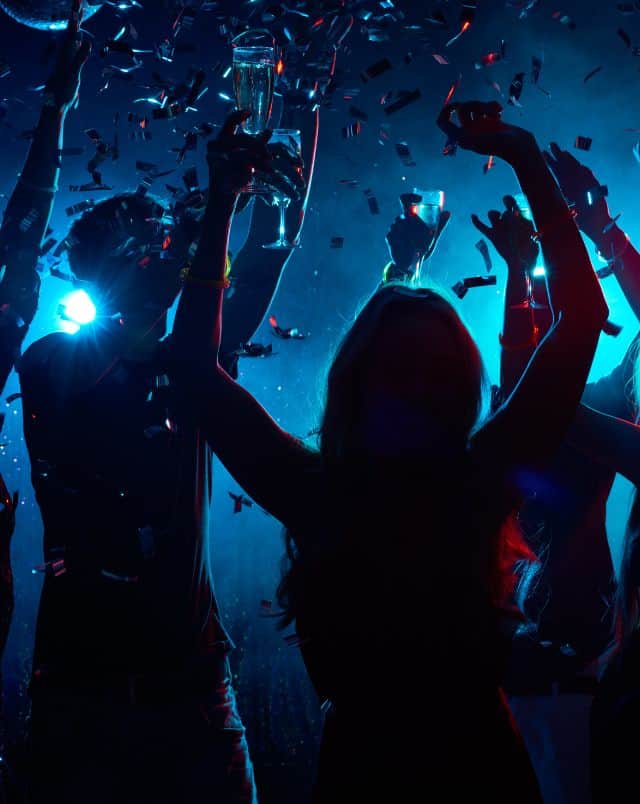 Close up shot of several people holding drinks in a club with low lights raising their arms as confetti falls