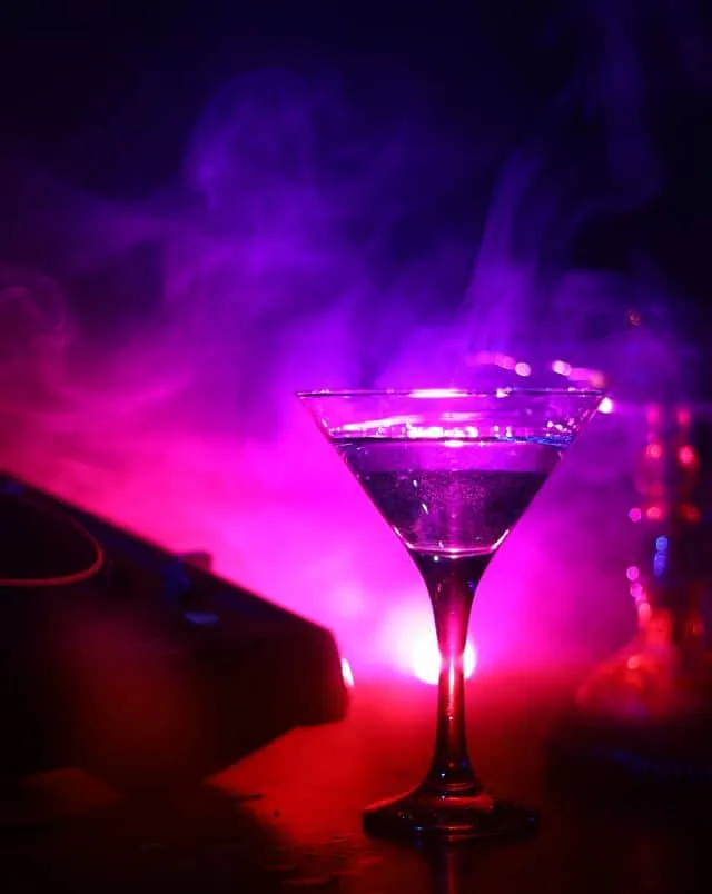 Close up shot of a drink in a martini glass surrounded by swirling smoke lit up by pink and red lights