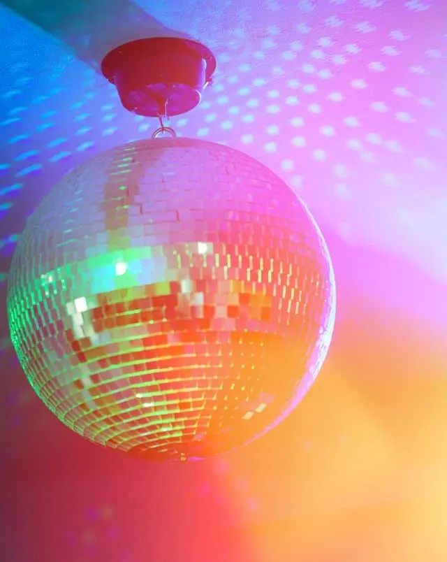 Close up shot of a silver disco ball suspended from the ceiling and bathed in a rainbow of colourful lights