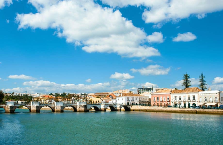 Amazing things to do in Tavira Portugal, Bridge crossing a body of green water next to residential townhouse buildings under a wide open blue sky with white fluffy clouds