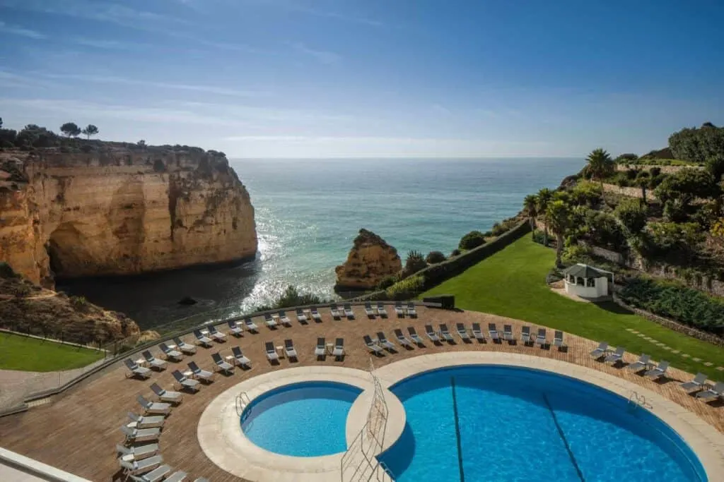 Try some of the greatest 5-star hotels Algarve Portugal can provide, view looking down onto outdoor pool area with two circular pools surrounded by rings of sun loungers with green areas to one side and rocky coastal cliffs to the other with the wide open sea in between