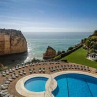 Try some of the greatest 5-star hotels Algarve Portugal can provide, view looking down onto outdoor pool area with two circular pools surrounded by rings of sun loungers with green areas to one side and rocky coastal cliffs to the other with the wide open sea in between