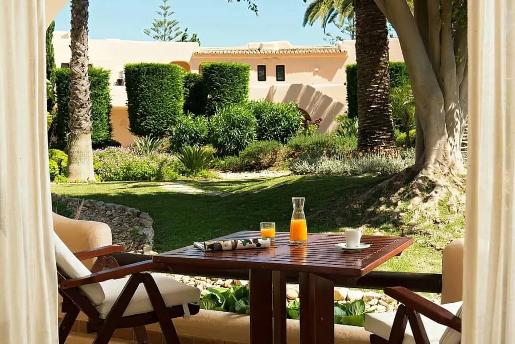 Find your favourite 5-star hotels in the Algarve, Portugal, view of outdoor breakfast area with wooden chairs and a table with orange juice and coffee on top with a green garden area behind