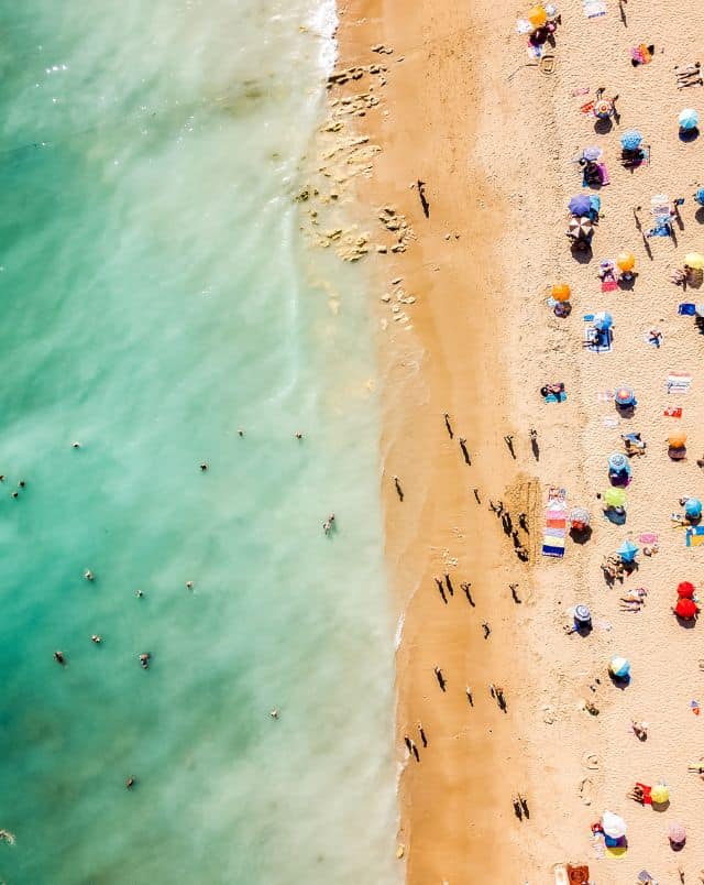 Aerial top down view of golden sandy beach with colourful beach umbrellas and sunbathers dotted around next to turquoise green sea water with people swimming in the shallows
