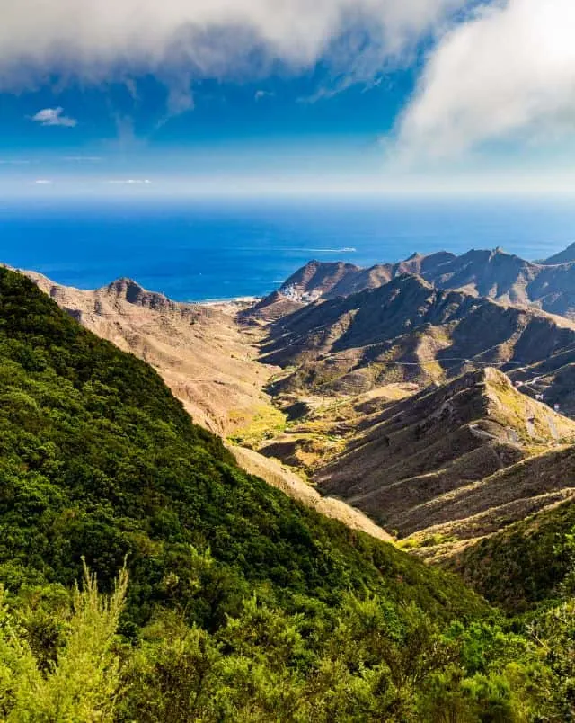 View of rolling hills and mountains leading towards the wide open sea with green grassy area in the foreground all under a bright blue sky with wispy clouds