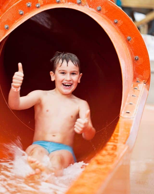 Visit the water parks in Costa Adeje Tenerife, Child smiling and giving two thumbs up while sliding down large water slide at a water park