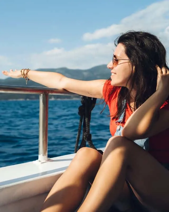 things to see and do in Silves Portugal, Smiling person in sunglasses sitting on the side of a boat holding their arm out over the edge of the railing with the sea behind and some tall rolling hills in the distance