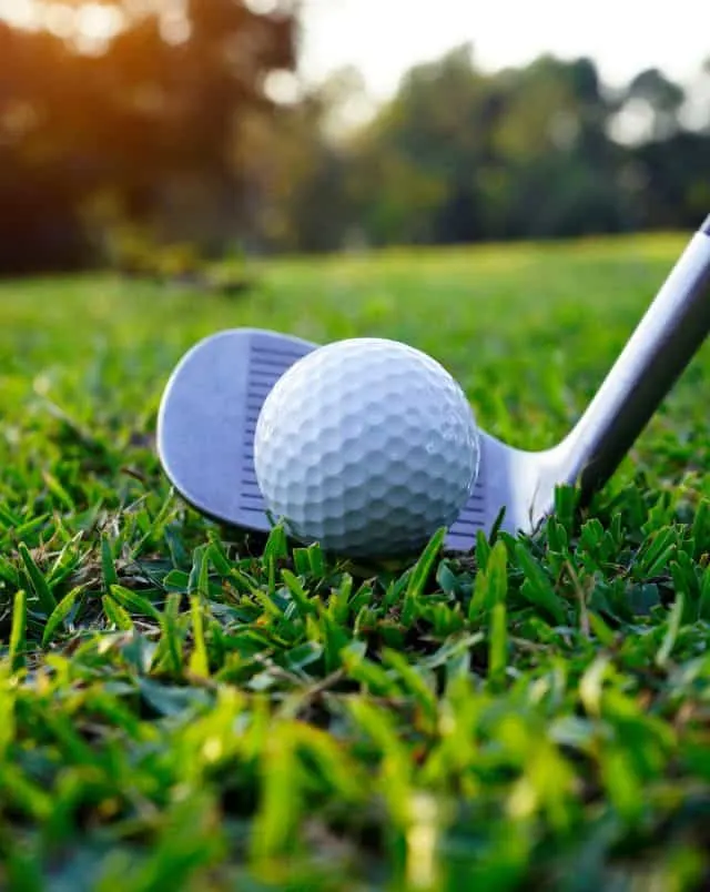 top activities in Vilamoura, Close up shot of a white golf ball sitting on some freshly cut green grass with a metal golf club poised for a shot behind