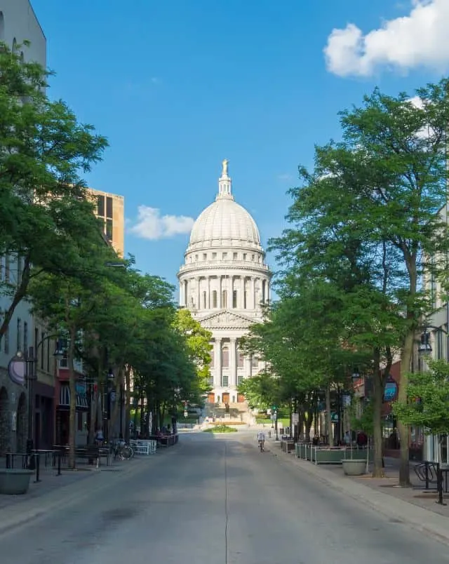 getaways in Wisconsin in March, View looking along an empty tree-lined street of a large white stone building with columns on the front and a large pointed dome on top under a bright blue sky with occasional white fluffy clouds