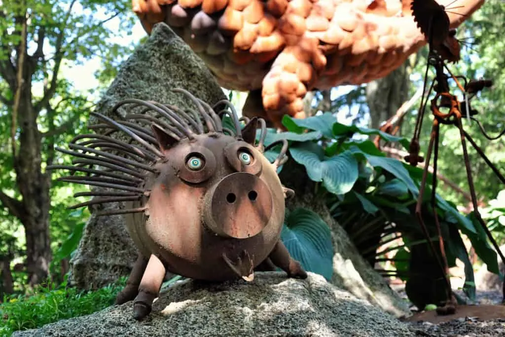 Close up shot of a metal sculpture of a porcupine standing on a small mound with a larger statue behind all surrounded by green trees on a sunny day
