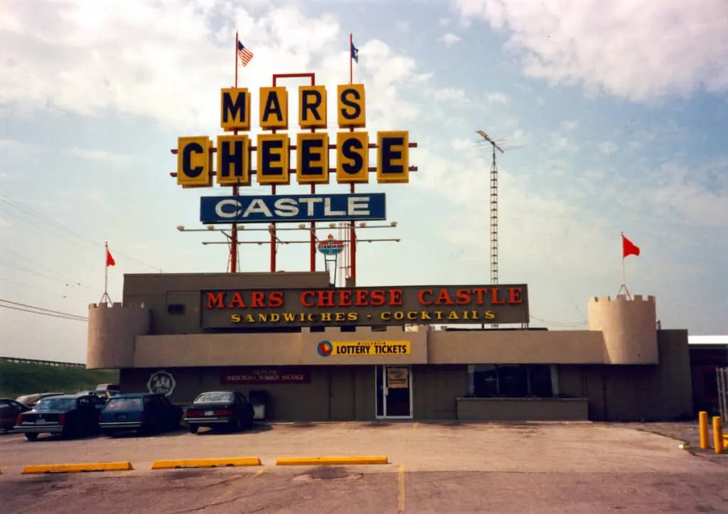 winter getaway ideas from Milwaukee, View of the front of the Mars Cheese Castle with two large signs and various flags on poles with a parking lot out front under a cloudy blue sky