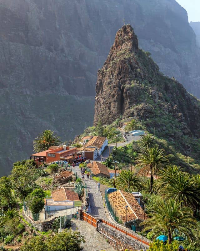 View of stone path running along ridge of mountain between palm trees and small buildings towards a prominent rocky peak covered in sporadic grass with a dramatic backdrop of a tall rocky mountainside