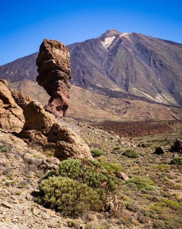 View of grassy dusty valley with large freestanding rock formation to one side and a large rocky mountain way off in the distance under a clear blue sky in mount teide, tenerife