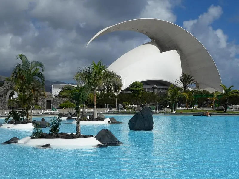 why visit the waterparks in Tenerife, View of large outdoor pool area with small islands and palm trees in front of a large modern building under a cloudy blue sky