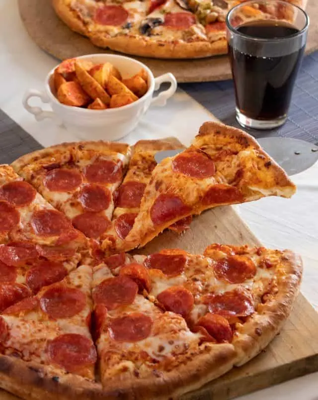 close up shot of a spatula retrieving a delicious slice of pepperoni pizza from a wooden board with a small bowl of chunky potato wedges and a glass of cola behind