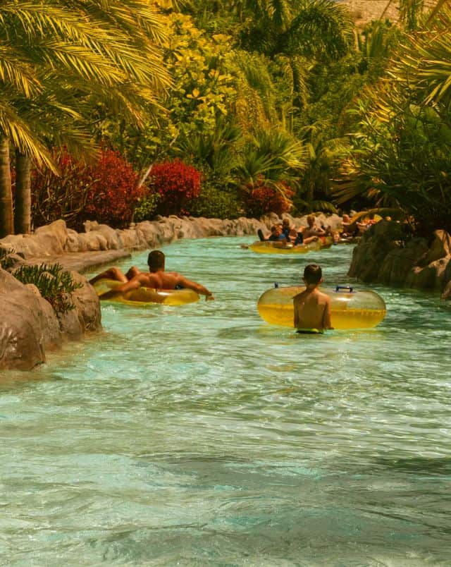 why visit Siam water park, Two people with inflatable water rings floating in a long river lined with stone paths and green trees with other water park visitors further downstream
