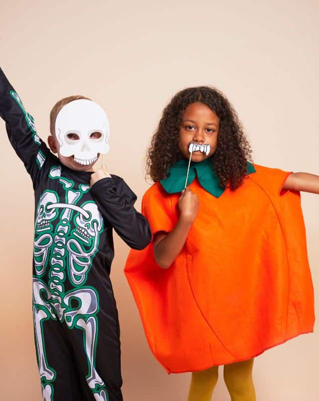 Halloween activities in Milwaukee for kids, two kids in Halloween costumes, a little white boy in a skeleton costume with a skull mask and a little black girl in a pumpkin costume holding up monster teeth to her mouth