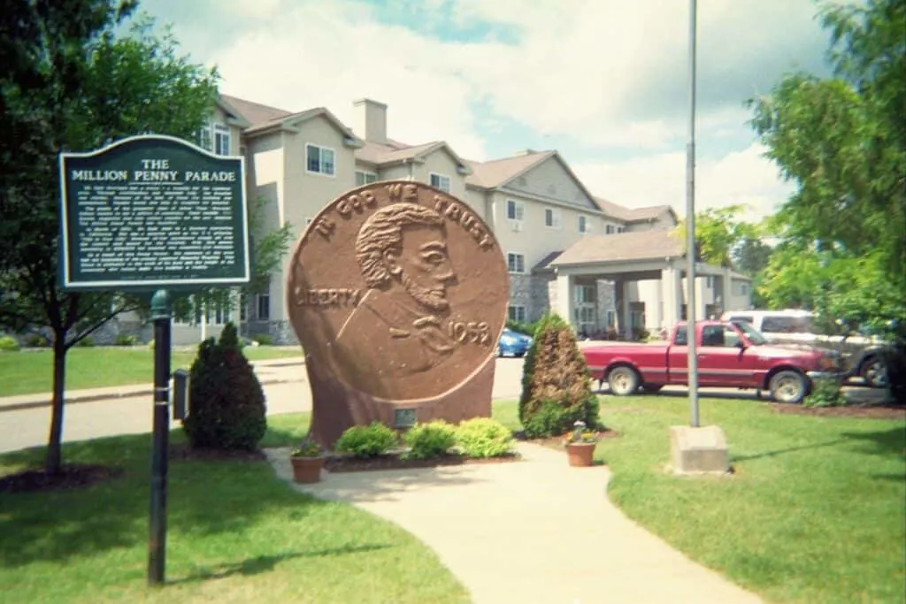 weird roadside attractions Wisconsin offers, View of the world's largest penny sitting outside in a small park area with a sign and flagpole nearby with some parked cars and residential houses behind