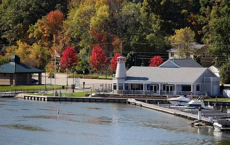 Wisconsin weekend getaways for families, View of Appleton Yacht Club with building and jetty with yachts moored in neat rows next to a large body of water with a thick canopy of trees behind in fall colors