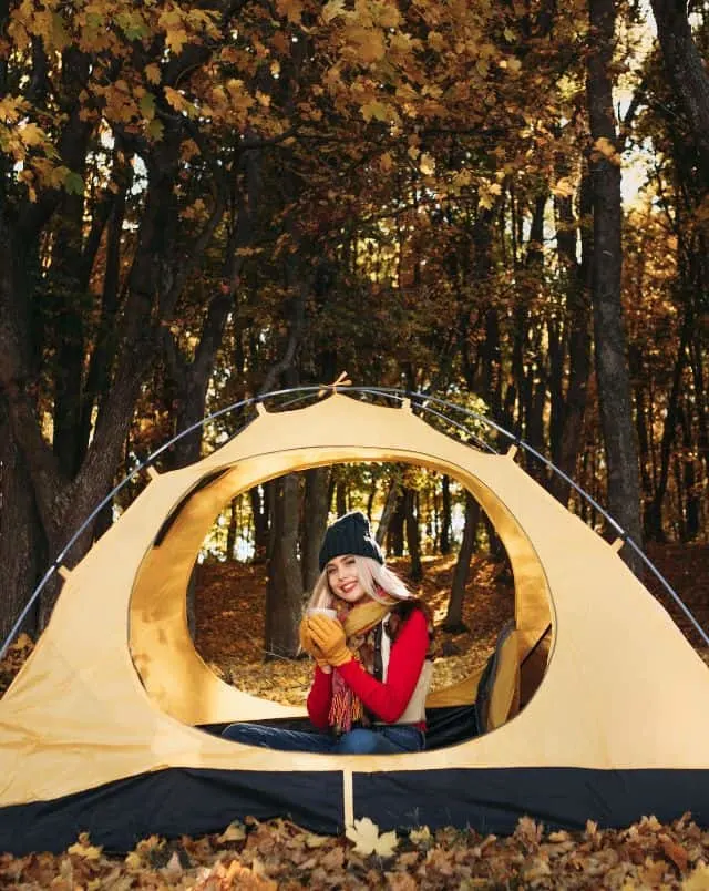 Door County things to do in September, Smiling person wearing winter clothing sitting with a hot drink inside a one-person tent in a forest full of fall-colored leaves and trees