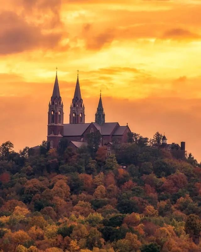 Ornate church with several pointed towers sitting atop a large hill covered in a forest of trees in different fall colors under a bright orange cloudy sky