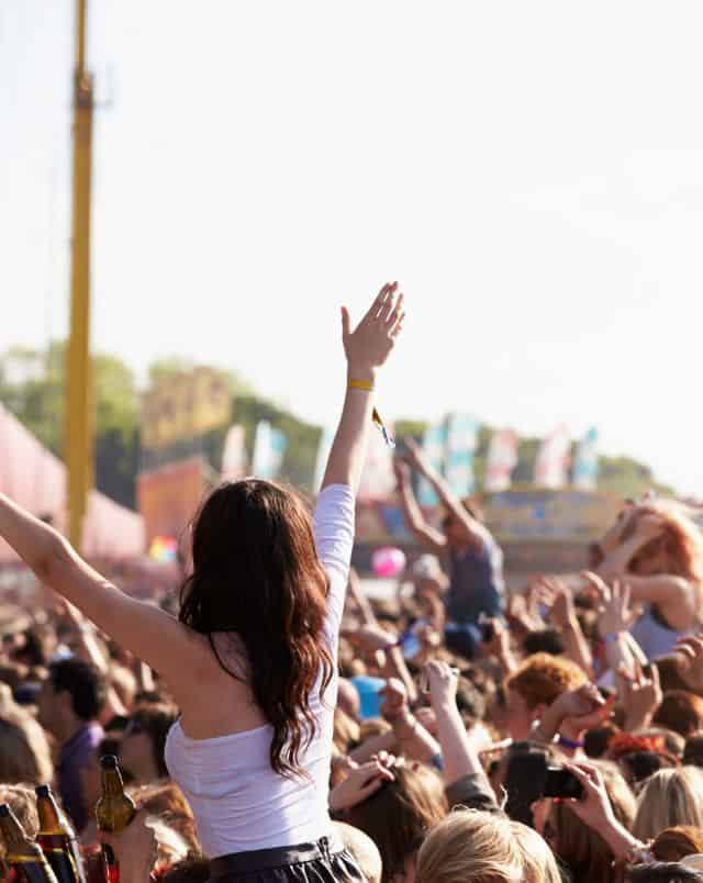 View from the back of a crowd of people appreciating some kind of spectacle off to the side with some spectators sitting on the shoulders of others and many people raising their arms in enjoyment and celebration on a bright clear day