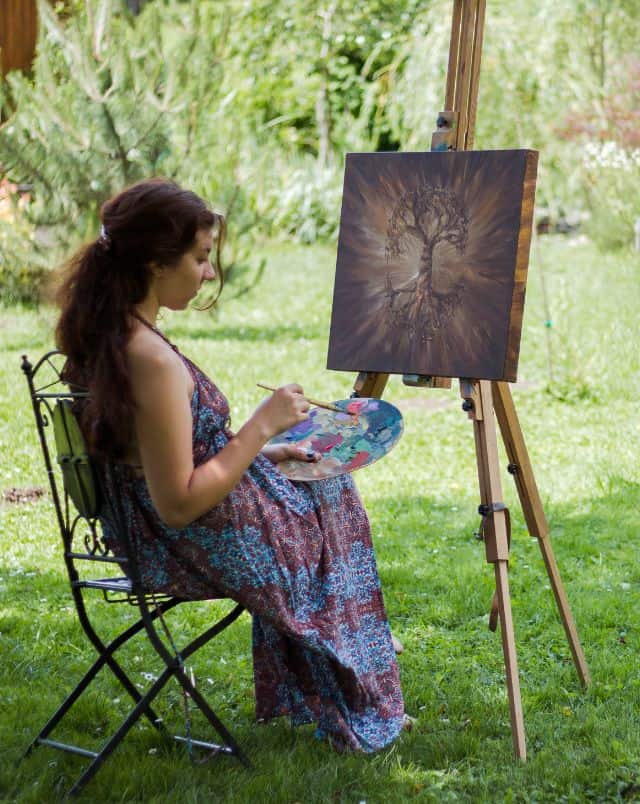 Enjoy September in Door County, Person sitting on a grassy lawn with a paintbrush and a palette in front of a canvas depicting painted artwork of a tree resting on a wooden easel