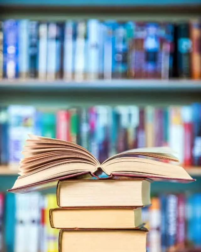 Close up shot of a stack of books with one book open on the top with a full bookshelf visible behind, photography coffee table books