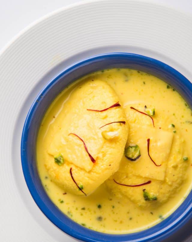 where to get vegan food in Milwaukee, Overhead shot of some ras malai with a creamy yellow sauce with green herb seasoning as well as some small strands of saffron sitting in a blue ceramic bowl