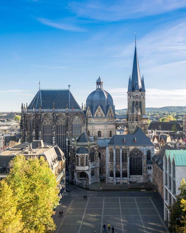 Discover where to travel from Luxembourg on a day trip, aerial view of a large cathedral with many stone sections topped with a variety of towers in different shapes with a small open square in front flanked by smaller modern buildings and green trees all under a bright sky with clouds
