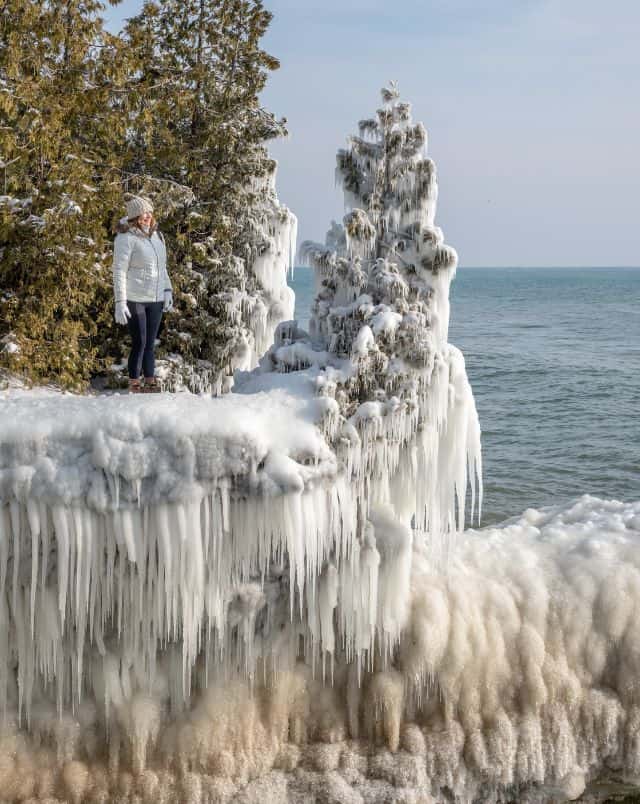 what to do in Sturgeon Bay in winter, Person standing by the coast in extremely cold weather next to a small tree and rock formation which have been covered in seawater which has frozen solid and is covered with many icicles