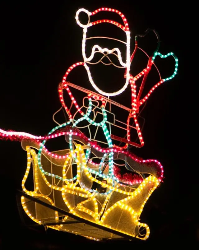 View from the street of a large neon sign shaped like Santa Claus waving and smiling from his golden sleigh against a pitch black night sky